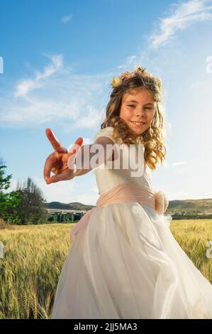 Lächelndes Mädchen in Kommunionkleid gestikulierenden Frieden Zeichen auf Feld Stockfoto