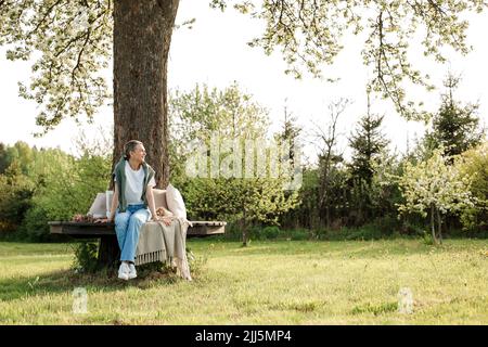 Lächelnde Frau, die auf einer Bank um den Baumstamm im Garten sitzt Stockfoto