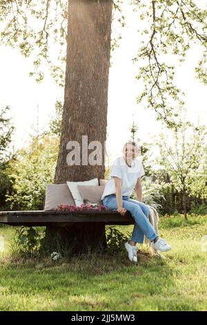 Glückliche Frau, die auf einer Bank um den Baumstamm im Garten sitzt Stockfoto