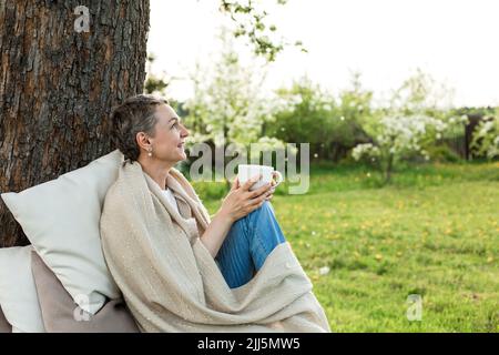 Glückliche Frau in eine Decke gewickelt, die einen Becher auf einer Bank um den Baumstamm hält Stockfoto