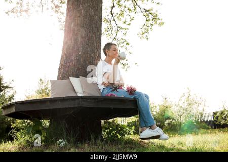 Lächelnde Frau mit Kaffeetasse, die auf einer Bank um den Baumstamm im Garten sitzt Stockfoto