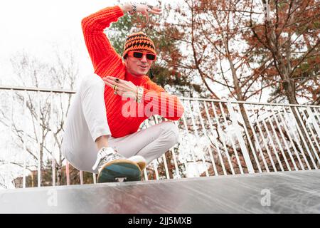 Lächelnder reifer Mann mit Strickmütze, der vor dem Geländer auf dem Skateboard hockt Stockfoto