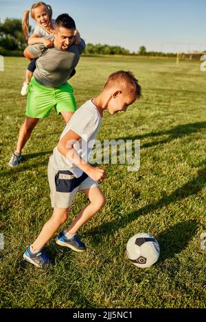 Junge spielt Fußball von Mann, der Mädchen auf dem Sportplatz mit dem Huckepack fährt Stockfoto