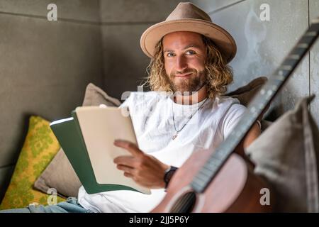 Lächelnder Gitarrist mit Buch und Gitarre zu Hause Stockfoto