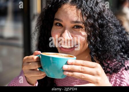 Glückliche junge Frau mit lockigen Haaren hält Kaffeetasse an sonnigen Tag Stockfoto