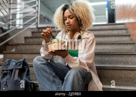 Junge Frau, die Salat isst, sitzt auf einer Treppe Stockfoto