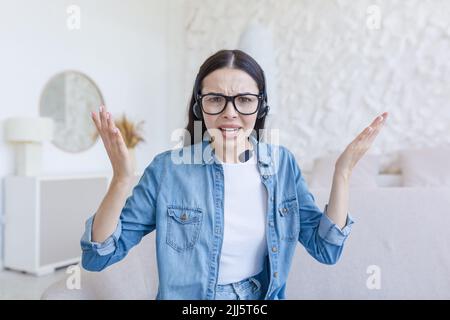Schöne Frau, die die Webcam anschaut, nervös und mit dem Gesprächspartner streitet, Headset für Videoanruf verwendet, zu Hause auf dem Sofa sitzt, Online-Konferenz und Studium Stockfoto