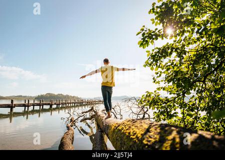 Mann mit ausgestreckten Armen balanciert auf einem umgestürzten Baum am See Stockfoto