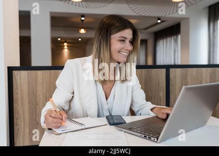 Lächelnde Geschäftsfrau, die auf den Laptop-Bildschirm schaut und im Restaurant Tagebuch schreibt Stockfoto