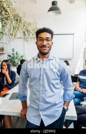 Lächelnder Geschäftsmann mit Kollegen im Hintergrund am Arbeitsplatz Stockfoto