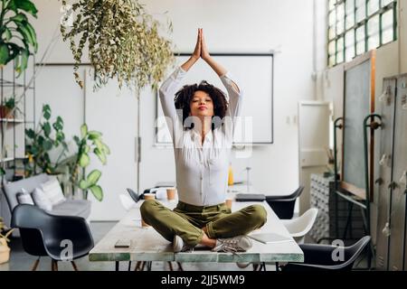 Junge Geschäftsfrau, die im Büro Yoga macht und die Hände auf den Schreibtisch geklammt hat Stockfoto