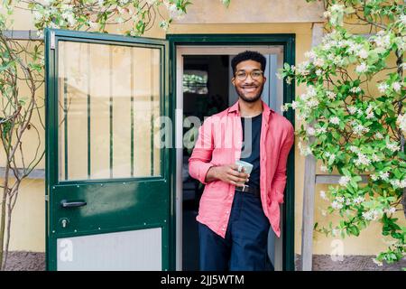 Lächelnder Geschäftsmann mit einer Tasse Kaffee an den Blumen vor der Tür Stockfoto