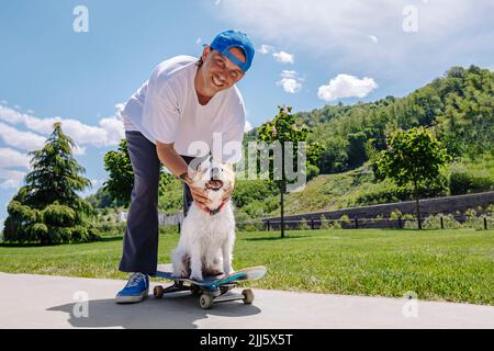 Glücklicher Mann mit Hund sitzt auf dem Skateboard im Park Stockfoto