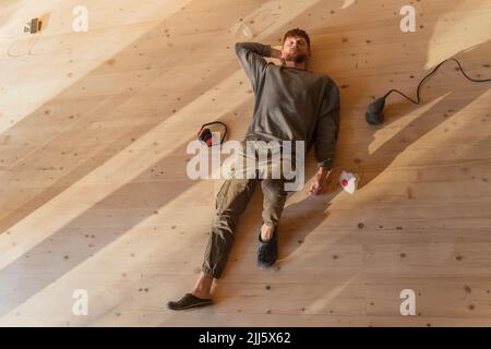 Mann, der nach der Arbeit auf dem frisch geschliffenen Holzboden liegt. Entspannen Sie sich unter der Sonne im Öko-Haus Stockfoto