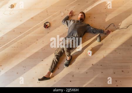 Mann, der nach der Arbeit auf dem frisch geschliffenen Holzboden liegt. Entspannen Sie sich unter der Sonne im Öko-Haus Stockfoto