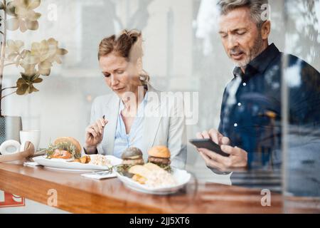Geschäftsfrau beim Mittagessen sitzend vom Geschäftsmann mit Smartphone im Café durch Glas gesehen Stockfoto