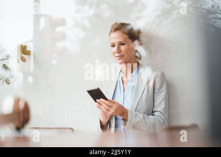 Geschäftsfrau, die im Café ein Smartphone benutzt, das durch Glas gesehen wird Stockfoto