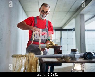 Klempner, der die Hanfdichtung an das Metallrohr am Arbeitsplatz bindet Stockfoto