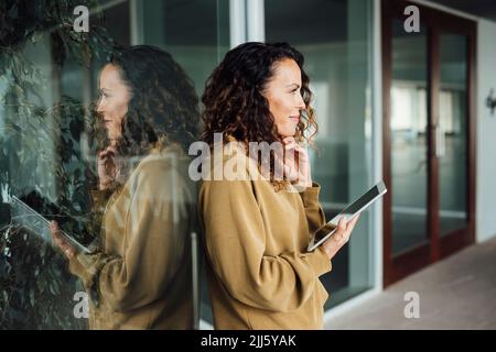 Lächelnde Geschäftsfrau mit Tablet-PC an Glaswand Stockfoto