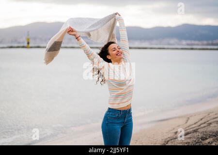 Fröhliche Frau mit erhobenen Armen hält Schal am Strand Stockfoto