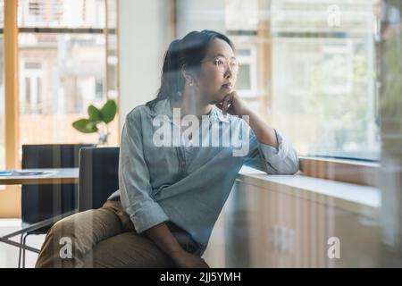 Geschäftsfrau mit der Hand auf dem Kinn durch das Glas des Büros gesehen Stockfoto