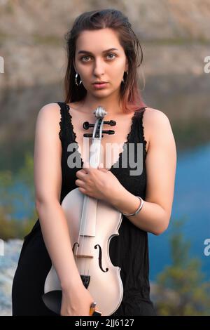 Schönes Mädchen mit einer Geige in den Händen am Ufer eines Bergsees Stockfoto