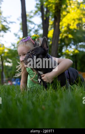 Ein kleines Mädchen versteckte sich hinter einer französischen Bulldogge, sie umarmte ihn und zusammen sehen sie gut zur Seite aus Stockfoto