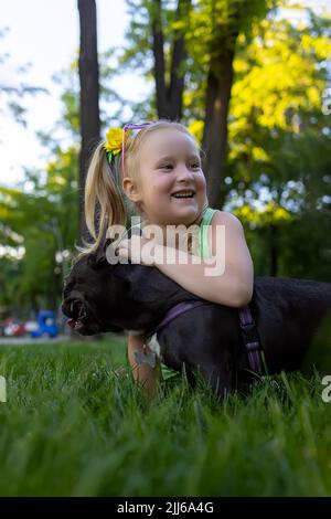 Ein Mädchen spielt im Park mit einem französischen Bulldoggen, der ihn umarmt und fröhlich lacht Stockfoto