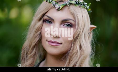 Porträt einer jungen Frau in Cosplay-Elf-Kleidung mit Make-up auf grünem Hintergrund. Fantastischer Look, langes blondes Haar, Waldkrone. Halloween-Konzept. Stockfoto