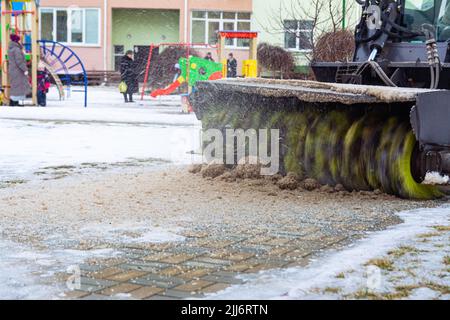 Ein Schneepflug entfernt Schnee vom Bürgersteig im Innenhof eines Wohnhauses. Im Winter reinigt eine rotierende Bürste den Straßenbelag von Schnee. Stockfoto