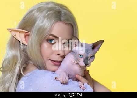 Porträt einer jungen Frau, die die Sphinx-Katze in der Nähe der Wange hält. Elfe im sommerblauen Kleid, mit schönen blonden lockigen langen Haaren Stockfoto