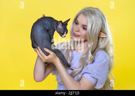 Junge blonde Cosplay-Elfe in blauem Sommerkleid mit schönen lockigen langen Haaren und Perlen im Ohr, die Sphinx Kätzchen in den Händen halten und lächelt und schaut Stockfoto