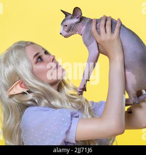 Cosplayer Elf junge blonde Frau im blauen Sommerkleid mit schönen lockigen langen Haaren und Perlen im Ohr sieht Sphinx Kitten liebevoll an Stockfoto