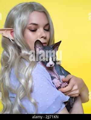 Nahaufnahme eines Cosplay-Elfen-jungen Weibchen mit Sphynx-Katze auf gelbem Hintergrund. Elfe in blauem Kleid, mit blondem welligen langen Haaren, durchbohrtes Ohr sieht Katze an Stockfoto