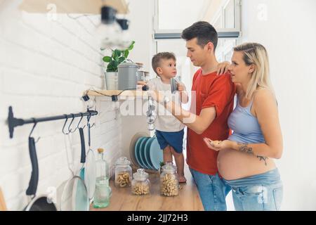 Glückliche Familie in der Küche. Die Schwangerin und ihr Partner zeigen ihrem Kindergartenksohn verschiedene Gewürze auf dem Küchenregal. Hochwertige Fotos Stockfoto