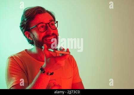 Junger 30 Jahre alter bärtiger Mann, der über das Smartphone mit dem Lautsprecher spricht, moderne Neonlichter. Hochwertige Fotos Stockfoto
