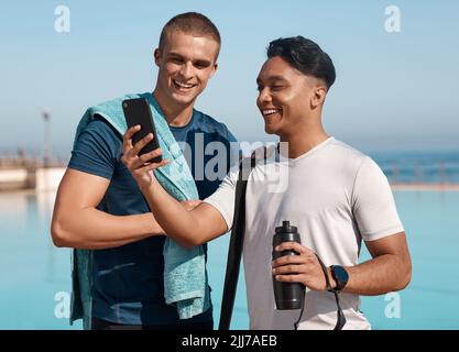Das Leben ist besser, wo es nasser ist. Zwei junge Männer benutzen ein Smartphone, bevor sie schwimmen gehen. Stockfoto