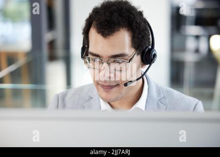 Lassen Sie mich sehen, was ich für Sie tun kann: Ein junger Callcenter-Agent, der an einem Computer in einem Büro arbeitet. Stockfoto