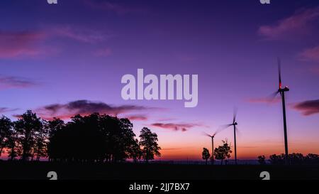 Die Silhouette von drei modernen Windmühlen in einem bunten Sonnenuntergang, neben einer Gruppe von Bäumen am Horizont, am Rande von Kiyú, San José, Uruguay Stockfoto