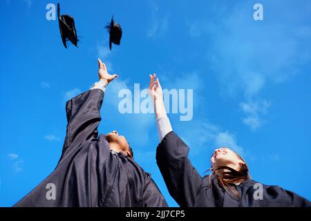 Nichts kann uns davon abhalten, Größe zu erreichen. Low-Angle-Aufnahme von zwei jungen Studenten, die ihre Hüte am Abschlusstag in die Luft werfen. Stockfoto