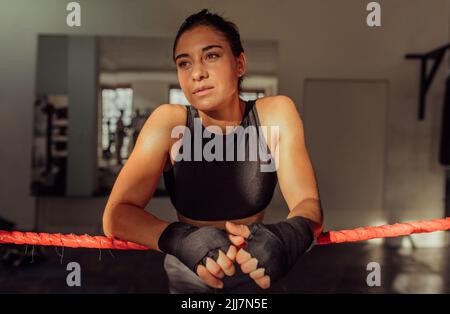 Die junge Boxerin schaut nachdenklich weg, während sie sich in einem Boxring an den Seilen lehnt. Sportliche junge Frau trainiert im Fitnessstudio. Stockfoto