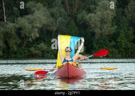 Ein Mädchen und ein Mann segeln in einem Kanu entlang des Dnjepr Flusses in der Stadt Dnepr in der Ukraine bei Sonnenuntergang mit der Flagge der Ukraine während des Krieges auf t Stockfoto