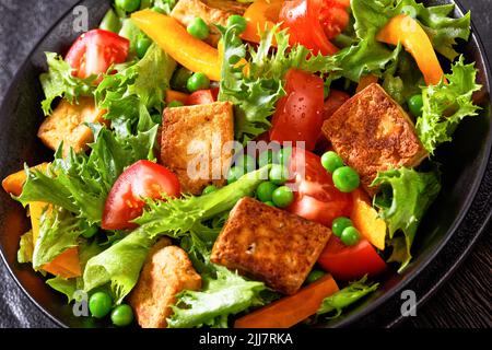 Gebratener Tofu-Salat mit knackigen Salatblättern, grünen Erbsen, Tomaten, gelbem Paprika in schwarzer Schüssel, Nahaufnahme Stockfoto