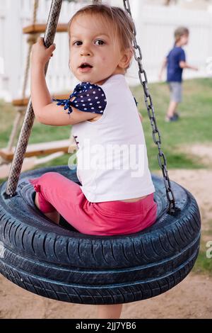 Kind schwingt auf den Ringen, schwingt von den Rädern auf dem Spielplatz. Mädchen, die Spaß während des Spaziergangs. Stark, sportlich und Füße im Sommer Stockfoto