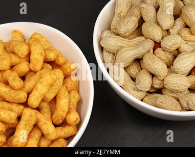 Portion Erdnussflips und natürlich geröstete Inshell-Erdnüsse in weißen Schalen auf schwarzem Hintergrund, Snack-Vergleich von Erdnüssen Stockfoto