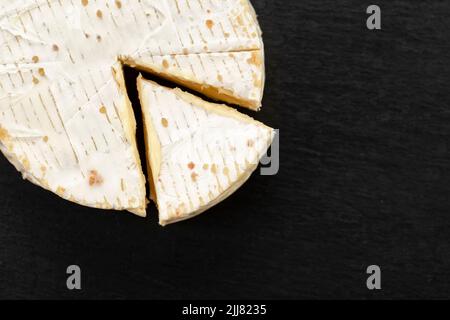 Brie-Käsesorte. Camembert-Käse. Frischer Brie-Käse und eine Scheibe auf einem schwarzen Brett. Italienischer, französischer Käse. Draufsicht. Stockfoto