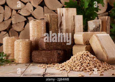 Verschiedene komprimierte Biokraftstoff-Briketts und Pyramide von Holzpellets auf Holztisch gegen Stapel von Brennholz bei Tageslicht angeordnet Stockfoto