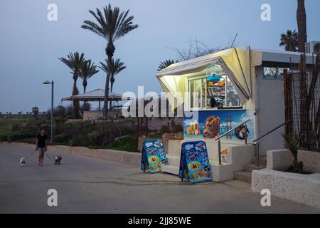 Eine Frau geht mit ihren Hunden in der Nähe eines Einkaufsbudes, der Snacks verkauft, auf einem Strandboulevard in Jaffa, Tel Aviv, Israel. Stockfoto