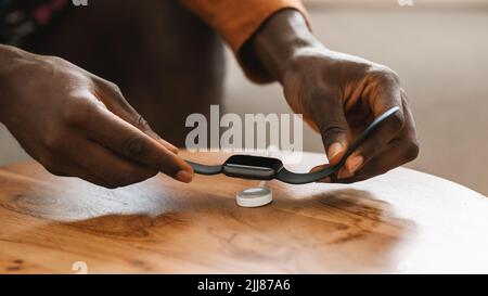 afroamerikanischer Mann, der die Smartwatch zum Aufladen auf einem kabellosen Ladegerät auf den Tisch legt, Nahaufnahme, abgeschnitten Stockfoto