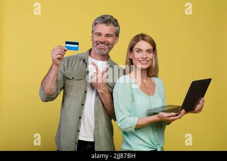 Online-Shopping-Konzept. Glückliche Ehegatten mittleren Alters mit Laptop und Kreditkarte, stehen auf gelbem Hintergrund Stockfoto